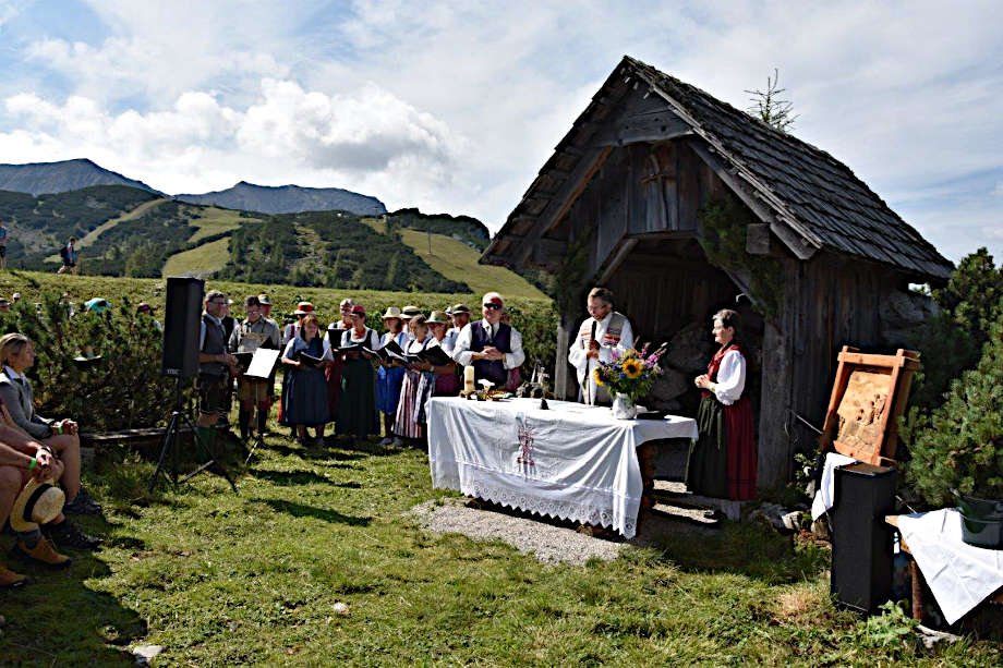 Feldmesse vor einem Stadel mit Pfarrer und Gästen, im Hintergrund die Alm bei schönem Wetter